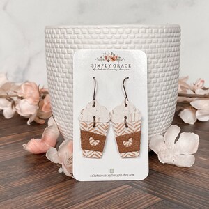 Puede incluir: Un par de pendientes de taza de caf&eacute; de color marr&oacute;n y blanco con un coraz&oacute;n recortado en la parte marr&oacute;n. Los pendientes est&aacute;n en una tarjeta blanca con el texto "SIMPLY GRACE by Dakota Country Designs" y la direcci&oacute;n del sitio web "dakotacountrydesigns.etsy.com".