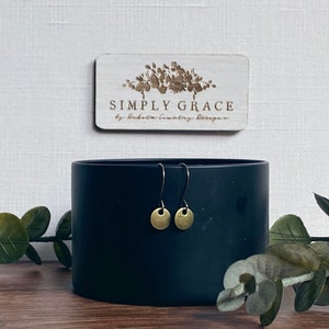 以下が含まれることがあります： シンプルなフックから小さな丸いディスクがぶら下がっている、ゴールドトーンのイヤリングのペア。イヤリングは黒い表面に表示されています。