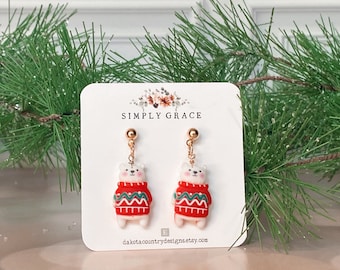 May include: A pair of white polar bear earrings wearing red and green sweaters. The earrings are on a white card with the text "SIMPLY GRACE" and "dakotacountrydesigns.etsy.com".