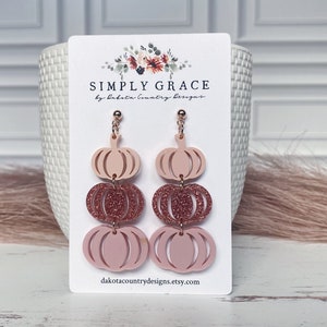 Può includere: Orecchini pendenti a forma di zucca rosa e oro rosa. Gli orecchini sono in acrilico e presentano tre zucche impilate. Le zucche superiore e inferiore sono rosa pieno, mentre la zucca centrale è glitter oro rosa. Gli orecchini sono attaccati a ganci dorati.