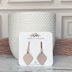 Puede incluir: Un par de pendientes de aro dorados con un diseño de hoja de cuero marrón claro. Los pendientes se muestran en una tarjeta blanca con el texto "SIMPLY GRACE" y una dirección de sitio web.