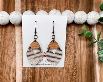 May include: A pair of dangle earrings with a geometric design. The earrings are made of wood and leather with a gold glitter accent. The earrings are hanging on a white card with the text "Country Designs Etsy" printed on it.