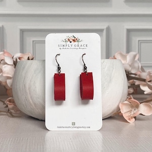 May include: Red leather earrings with a simple rectangular shape. The earrings are on a white card with the text "SIMPLY GRACE by Dakota Country Designs" and the website address "dakotacountrydesigns.etsy.com".