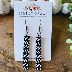 May include: A pair of black and white cork earrings with a geometric pattern. The earrings are hanging from gold hooks on a white card with the text "SIMPLY GRACE by Dakota Country Designs".
