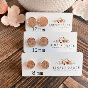 Pu&ograve; includere: Tre paia di orecchini a bottone dorati con una finitura marrone scintillante. Gli orecchini sono esposti su carte bianche con il testo "SIMPLY GRACE" e le dimensioni 12 mm, 10 mm e 8 mm.