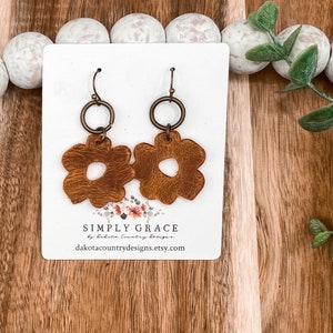 Pode incluir: Um par de brincos de couro marrom em forma de flor com um aro e um gancho dourados. Os brincos são exibidos em um cartão branco com o texto "SIMPLY GRACE by dakota country designs dakotacountrydesigns.etsy.com".