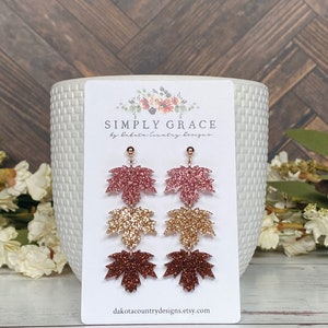 May include: A pair of dangle earrings featuring three layered maple leaves in shades of pink, gold, and brown glitter. The earrings are made of resin and have gold-toned hooks. The earrings are displayed on a white card with the text "SIMPLY GRACE by Dakota Country Designs" and "dakotacountrydesigns.etsy.com".