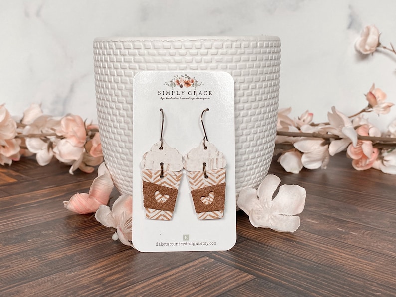Puede incluir: Un par de pendientes de taza de caf&eacute; de color marr&oacute;n y blanco con un coraz&oacute;n recortado en la parte marr&oacute;n. Los pendientes est&aacute;n en una tarjeta blanca con el texto "SIMPLY GRACE by Dakota Country Designs" y la direcci&oacute;n del sitio web "dakotacountrydesigns.etsy.com".