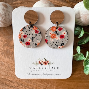 Puede incluir: Un par de pendientes de madera con un patrón floral. Los pendientes son redondos y tienen un fondo gris con flores naranjas, rojas y blancas. Los pendientes están unidos a un poste de madera.