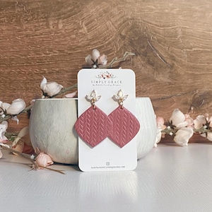 May include: A pair of pink, textured, diamond-shaped earrings with gold accents and clear rhinestones. The earrings are on a white card with the text "SIMPLY GRACE by Dakota Country Designs".