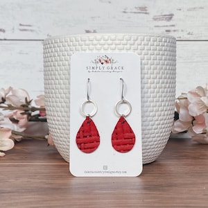 May include: A pair of red teardrop earrings with a woven texture, hanging from silver hoops. The earrings are displayed on a white card with the text "SIMPLY GRACE" and "dakotacountrydesigns.etsy.com". The background includes a white textured vase and pink flowers.