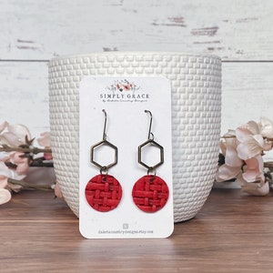May include: A pair of red leather earrings with a woven texture. The earrings feature a bronze-colored geometric hexagon shape and a circular red leather base. The earrings are displayed on a white card with the text "SIMPLY GRACE" and "dakotacountrydesigns.etsy.com".