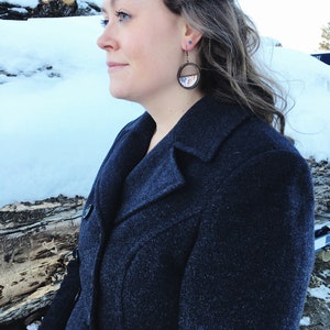 May include: A woman wearing a dark blue coat with a pair of round, wooden earrings with a colorful design. The earrings are hanging from her ears.