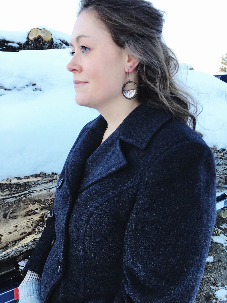 May include: A woman wearing a dark blue coat with a pair of round, wooden earrings with a colorful design. The earrings are hanging from her ears.