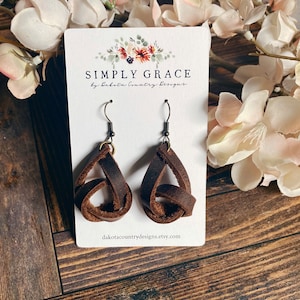 May include: Brown leather earrings with a knotted design. The earrings are on a white card with the text "SIMPLY GRACE by Dakota Country Designs" and the website "dakotacountrydesigns.etsy.com".