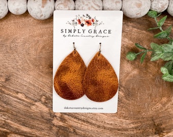 Peut inclure: Une paire de boucles d'oreilles en forme de goutte en cuir marron avec des crochets dorés. Les boucles d'oreilles sont présentées sur une carte blanche avec le texte "SIMPLY GRACE by Dakota Country Designs".