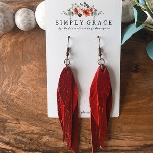 May include: Red leather earrings with a feather design. The earrings are on a white card with the text "SIMPLY GRACE by Dakota Country Designs".