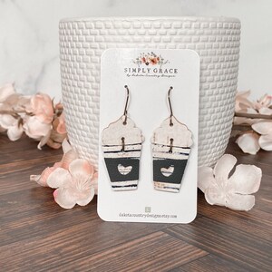 Puede incluir: Un par de pendientes de taza de caf&eacute; blanca y negra con un coraz&oacute;n recortado en una tarjeta blanca con el texto "SIMPLY GRACE by Dakota Country Designs" y el sitio web "dakotacountrydesigns.etsy.com".