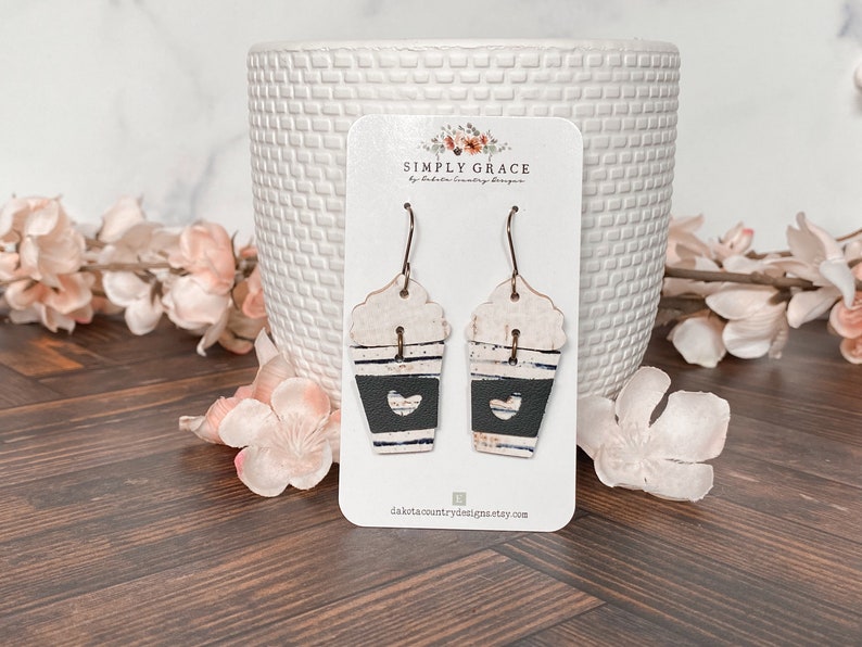 Puede incluir: Un par de pendientes de taza de caf&eacute; blanca y negra con un coraz&oacute;n recortado en una tarjeta blanca con el texto "SIMPLY GRACE by Dakota Country Designs" y el sitio web "dakotacountrydesigns.etsy.com".