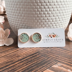 Peut inclure: Une paire de boucles d'oreilles clous dor&eacute;es avec un motif marbr&eacute; vert et brun. Les boucles d'oreilles sont pr&eacute;sent&eacute;es sur une carte blanche avec le texte "SIMPLY GRACE by Dakota Country Designs".