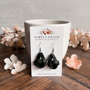 May include: Black leather earrings with a knotted design. The earrings are displayed on a white card with the text "SIMPLY GRACE by Dakota Country Designs" and the website address "dakotacountrydesigns.etsy.com".