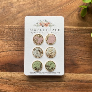 Peut inclure: Un ensemble de six boucles d'oreilles rondes sur une carte blanche. Les boucles d'oreilles ont des bordures dorées et présentent des motifs texturés en rose, blanc et vert avec des accents de feuilles d'or. La carte indique "SIMPLY GRACE" et "dakotacountrydesigns.etsy.com".