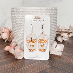 Puede incluir: Un par de pendientes colgantes con un dise&ntilde;o de taza de caf&eacute; blanco y naranja con un coraz&oacute;n recortado. Los pendientes est&aacute;n en una tarjeta blanca con el texto "SIMPLY GRACE by Dakota Country Designs" y la direcci&oacute;n del sitio web "dakotacountrydesigns.etsy.com".