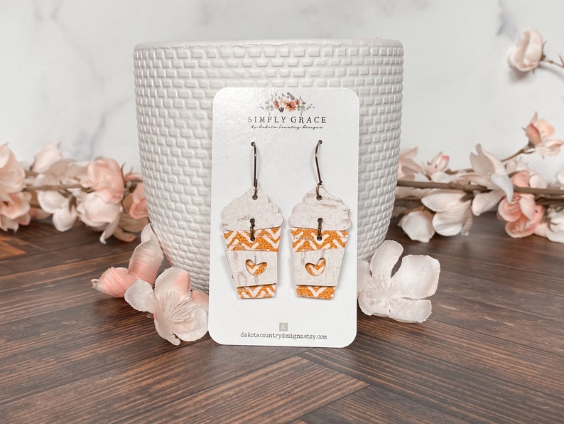 Puede incluir: Un par de pendientes colgantes con un dise&ntilde;o de taza de caf&eacute; blanco y naranja con un coraz&oacute;n recortado. Los pendientes est&aacute;n en una tarjeta blanca con el texto "SIMPLY GRACE by Dakota Country Designs" y la direcci&oacute;n del sitio web "dakotacountrydesigns.etsy.com".