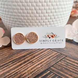 Peut inclure: Une paire de boucles d'oreilles clous dorées avec un motif de paillettes brunes. Les boucles d'oreilles sont présentées sur une carte blanche avec le texte "SIMPLY GRACE by babalu country design".