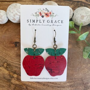 May include: Red and green cork apple earrings with gold hooks. The earrings are on a white card with the text "SIMPLY GRACE by Dakota Country Designs" and the website "dakotacountrydesigns.etsy.com".
