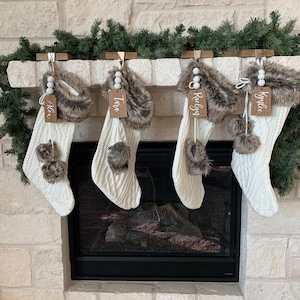May include: Four white knit stockings with faux fur trim and wooden name tags hanging on a fireplace mantel. The names on the tags are Alex, Tara, Raegan, and Kyler.