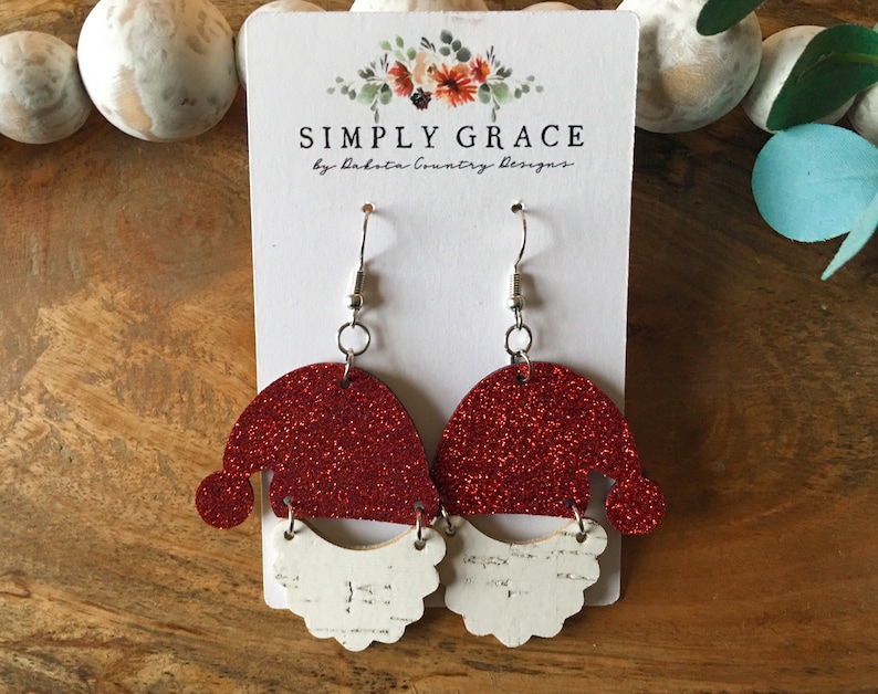 Pu&ograve; includere: Orecchini a forma di cappello di Babbo Natale con glitter rosso e barba di glitter bianco. Gli orecchini sono su una superficie di legno con un cartoncino bianco dietro. Il cartoncino dice "SIMPLY GRACE by Dakota Country Designs".