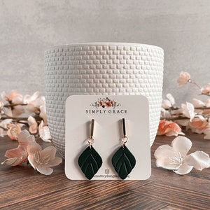 May include: A pair of dark green leaf-shaped dangle earrings with gold accents. The earrings are on a white card with the text "SIMPLY GRACE" and a website address.