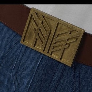 May include: A brown leather belt with a rectangular gold-coloured buckle. The buckle features a geometric design. The belt is over blue denim fabric.