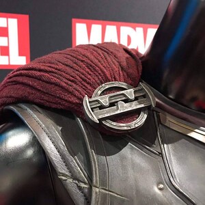 May include: Close-up of a costume shoulder piece. The shoulder armor is dark gray with a metallic sheen, and a burgundy fabric is draped over it. A silver-colored, circular clasp secures the fabric. The background features the word "MARVEL" in red and white.