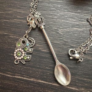 Könnte beinhalten: Silberfarbene Halskette mit einem Löffelanhänger mit Kronendetail und einem Elefanten-Charm. Der Elefanten-Charm ist mit grünen, rosa und türkisfarbenen Edelsteinen verziert. Die Halskette wird auf einer dunklen Holzoberfläche präsentiert.