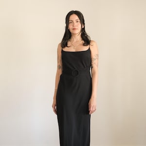 May include: A floor-length black dress featuring thin spaghetti straps, a straight neckline, and a wide fabric waistband accented with a circular ring detail at the center. The garment has a fluid, straight-cut skirt that falls to the floor.