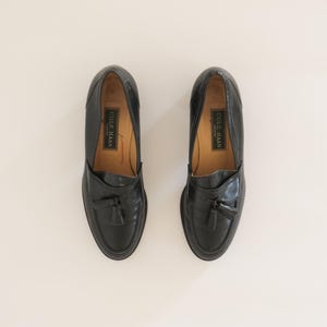Pu&ograve; includere: Un paio di mocassini in pelle nera con nappine e un'etichetta del marchio Cole Haan. I mocassini hanno una punta arrotondata e un tacco basso.