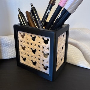 Peut inclure: Un porte-stylos noir avec un motif tissé et des découpes de Mickey Mouse. Le porte-stylos est rempli de stylos, dont des stylos bruns, roses et gris.