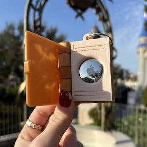 May include: An orange and white keychain shaped like a small book with a silver Apple logo inside. The keychain is open and the Apple logo is visible.