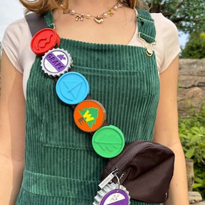 May include: A green corduroy overall dress with a brown fanny pack and a chain of colorful bottle caps with various designs. The bottle caps include a compass, a tree, a leaf, and a grape soda logo.