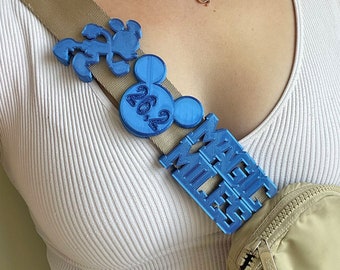 May include: A tan crossbody bag with a blue Mickey Mouse themed charm. The charm has the words "Magic Kingdom" and "2023" on it.