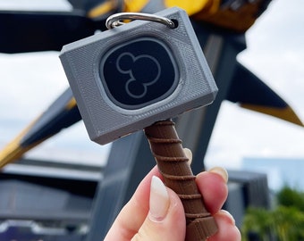 May include: A grey 3D printed keychain shaped like Thor's hammer with a black Mickey Mouse logo on the front. The keychain is being held by a hand with white nail polish.