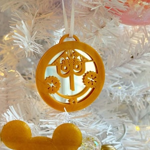 May include: A gold Christmas ornament shaped like the iconic Spaceship Earth ride at Epcot, a Disney theme park. The ornament is hanging on a white artificial Christmas tree.