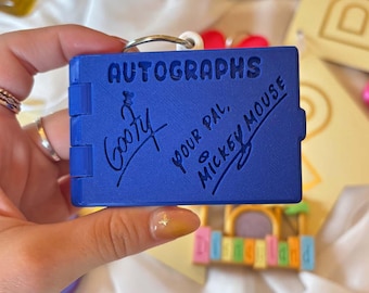 Könnte beinhalten: Ein blauer Schlüsselanhänger in Form eines Autogrammbuchs mit dem Wort "AUTOGRAPHS" auf der Vorderseite. Der Schlüsselanhänger hat Unterschriften von Goofy und Mickey Mouse. Der Schlüsselanhänger wird von einer Person gehalten.
