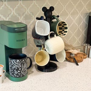 May include: A green Keurig coffee maker next to a black mug tree stand shaped like a mouse head. The stand holds several ceramic mugs with various patterns. A black mug with white text reading Happiest Place on Earth sits on the machine base.
