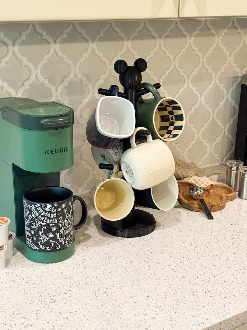 May include: A green Keurig coffee maker next to a black mug tree stand shaped like a mouse head. The stand holds several ceramic mugs with various patterns. A black mug with white text reading Happiest Place on Earth sits on the machine base.