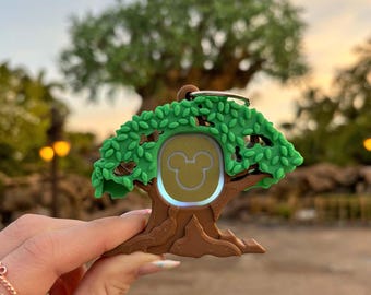 Könnte beinhalten: Ein grüner und brauner Plastik-Schlüsselanhänger in Form eines Baumes mit einem Mickey-Maus-Symbol in der Mitte. Der Schlüsselanhänger hat einen silbernen Ring zum Anbringen von Schlüsseln.
