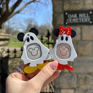Puede incluir: Dos llaveros blancos en forma de fantasma con orejas negras de Mickey Mouse y un lazo rojo en uno. El llavero tiene una cabeza gris de Mickey Mouse dentro del fantasma. Los llaveros se sostienen en una mano frente a una pared de piedra con un letrero que dice "Oak Hill Cemetery."