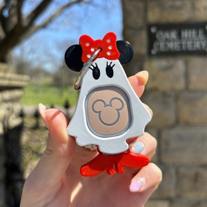 Puede incluir: Llavero blanco en forma de fantasma con un lazo rojo con lunares blancos, orejas negras y una cabeza de Mickey Mouse marr&oacute;n en la parte delantera. El llavero tiene un anillo plateado y patas rojas.